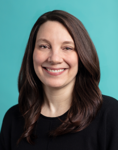 A person with long brown hair and a black shirt smiles against a turquoise background.